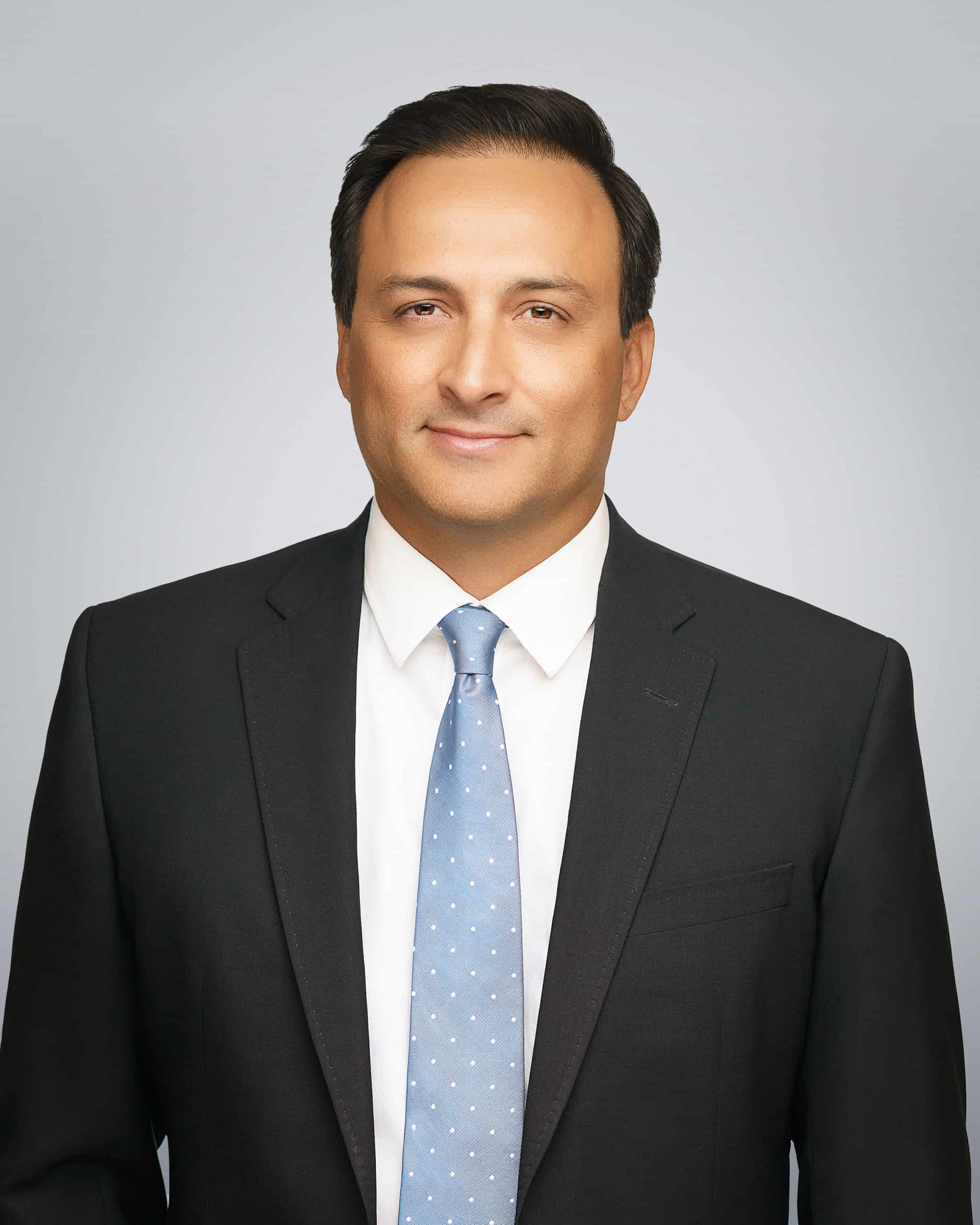 Attorney Doll Amir Eley, a professional lawyer, dressed in a black suit and light blue tie, smiling.